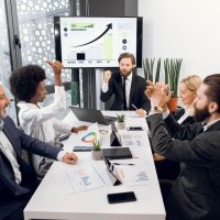 Smiling diverse cheerful multiracial businesspeople sit at office desk, celebrating successful project, gesturing, laughing, showing thumbs up, clapping hands and sharing business ideas together.